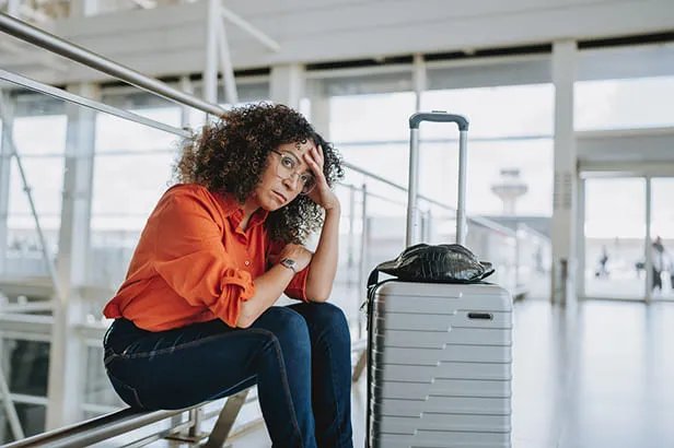Donna seduta in aeroporto con valigia grigia, sguardo preoccupato