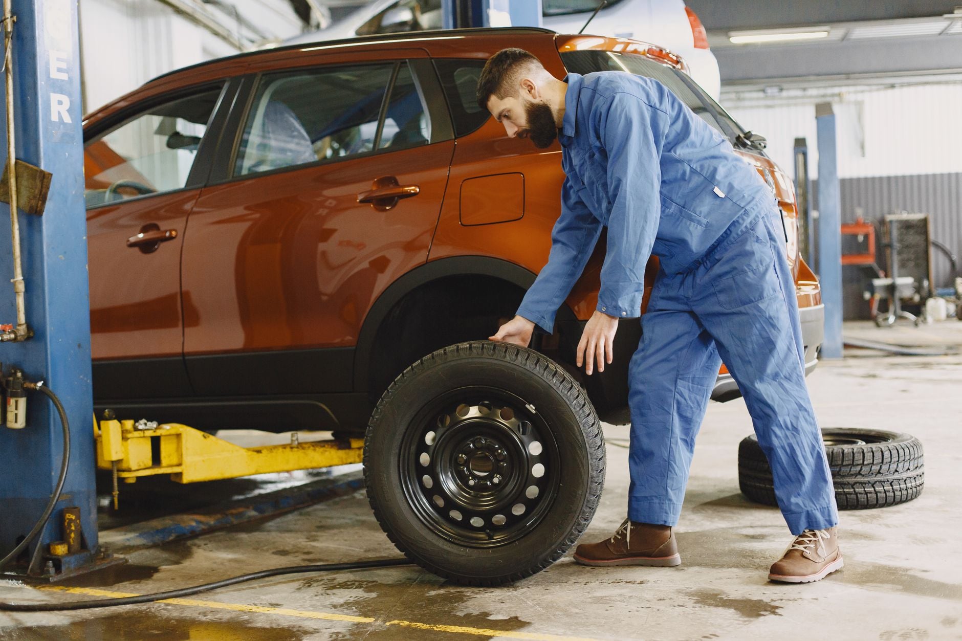 Mécanicien en combinaison bleue inspectant un pneu d'un SUV orange dans un atelier automobile.