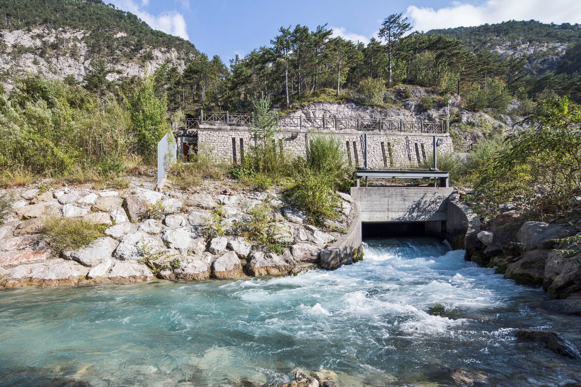 Diga in pietra con acqua azzurra tra montagne rocciose e vegetazione