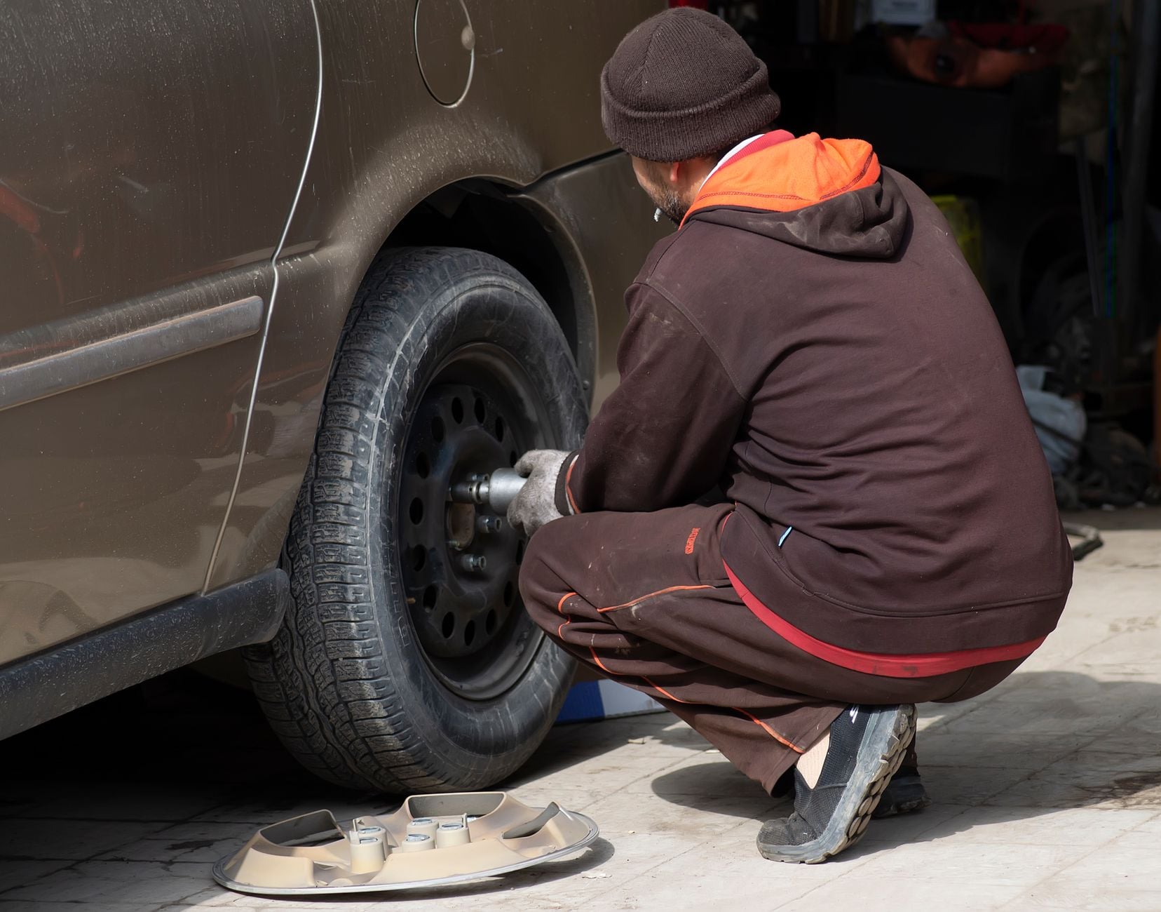 Homme en veste marron changeant un pneu de voiture avec une clé à tube, pièces de roue visibles au sol.