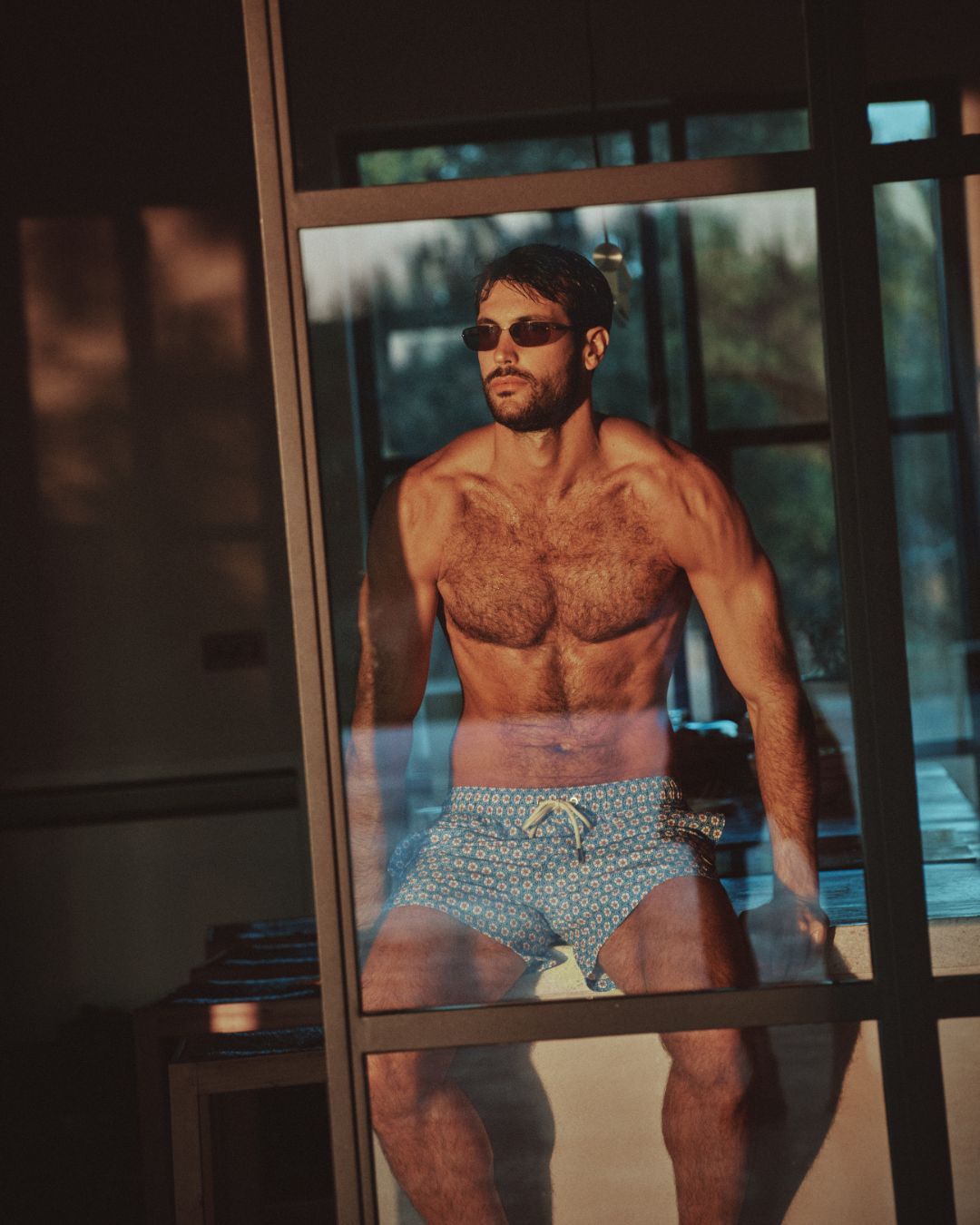 Uomo con occhiali da sole in posa davanti a una porta a vetri, indossa costume da bagno a fantasia