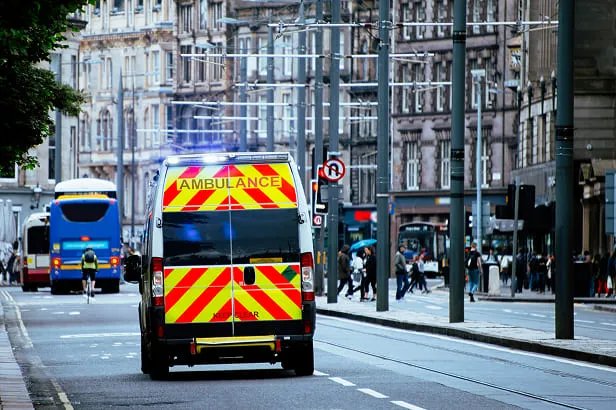 Ambulanza con luci blu accese in una strada cittadina affollata tra edifici