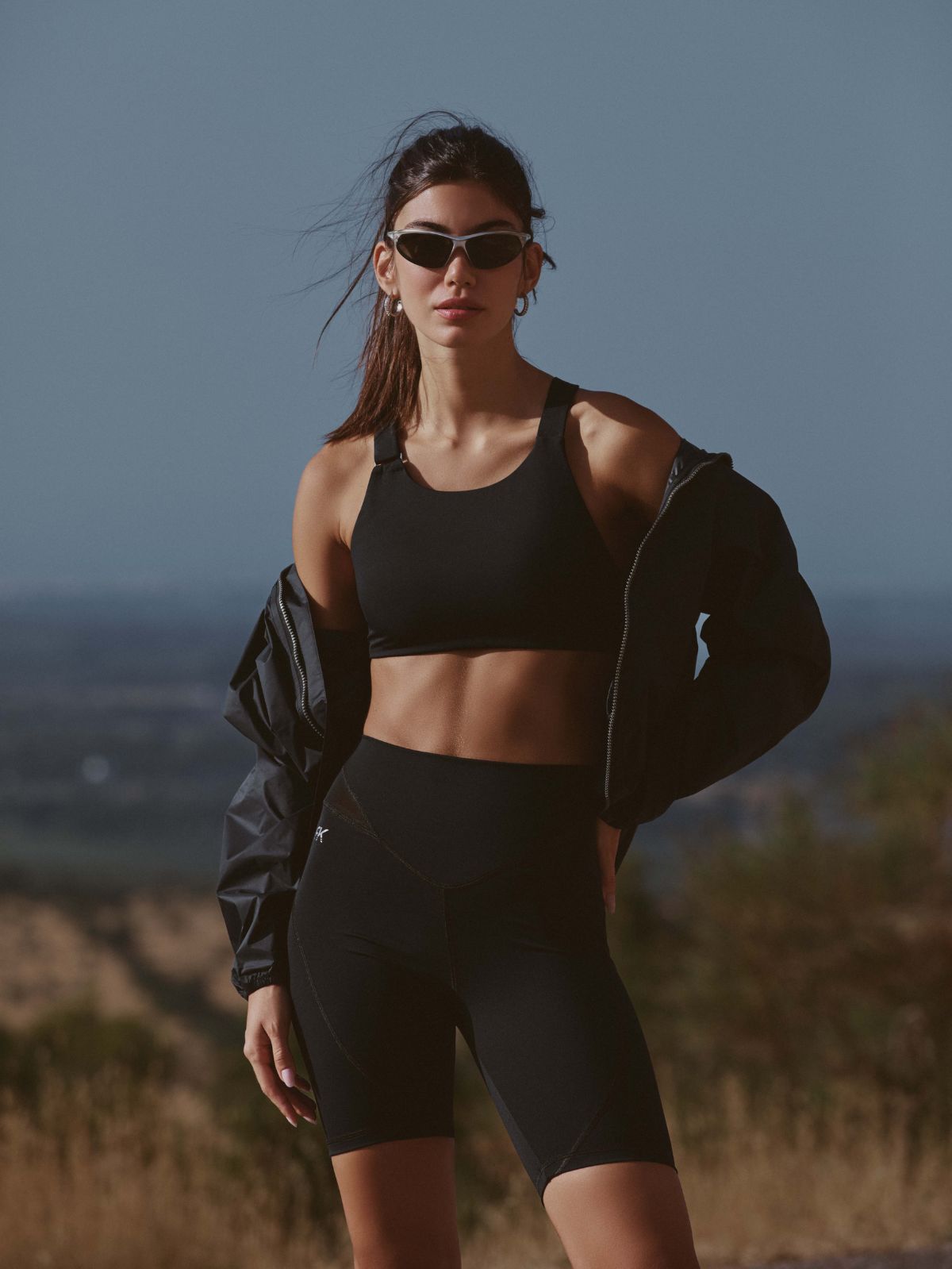 Donna in abbigliamento sportivo nero con giacca, occhiali da sole, posa in paesaggio collinare